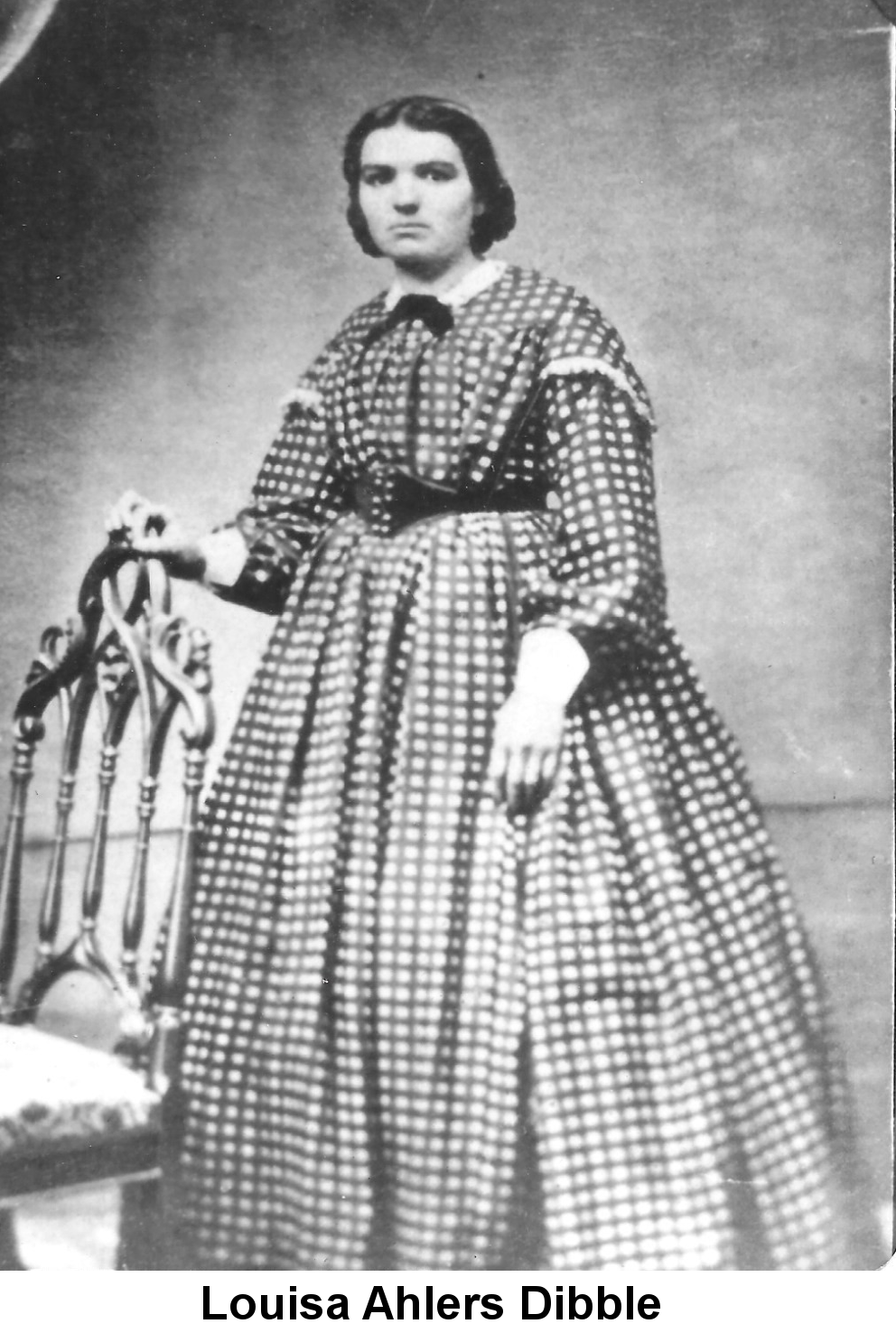 IMAGE/PHOTO: Louisa Ahlers Dibble: Black and white photo of a young woman in a checked hoop skirted-dress and short black hair standing with her right hand resting on the back of an ornate chair.